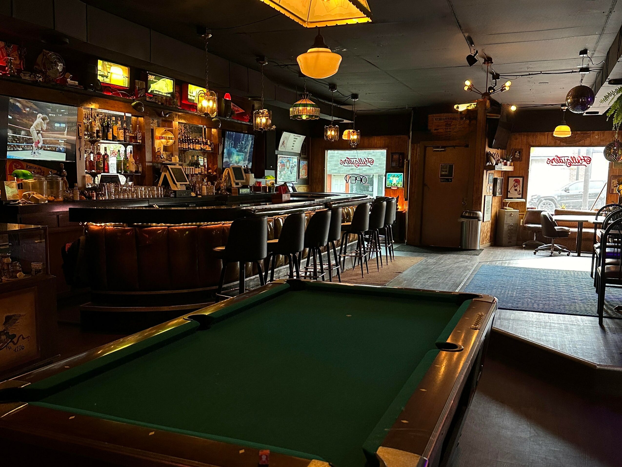 Bar with seating, pool table in foreground, dim lighting, TVs on wall, windows letting in natural light, cozy atmosphere.