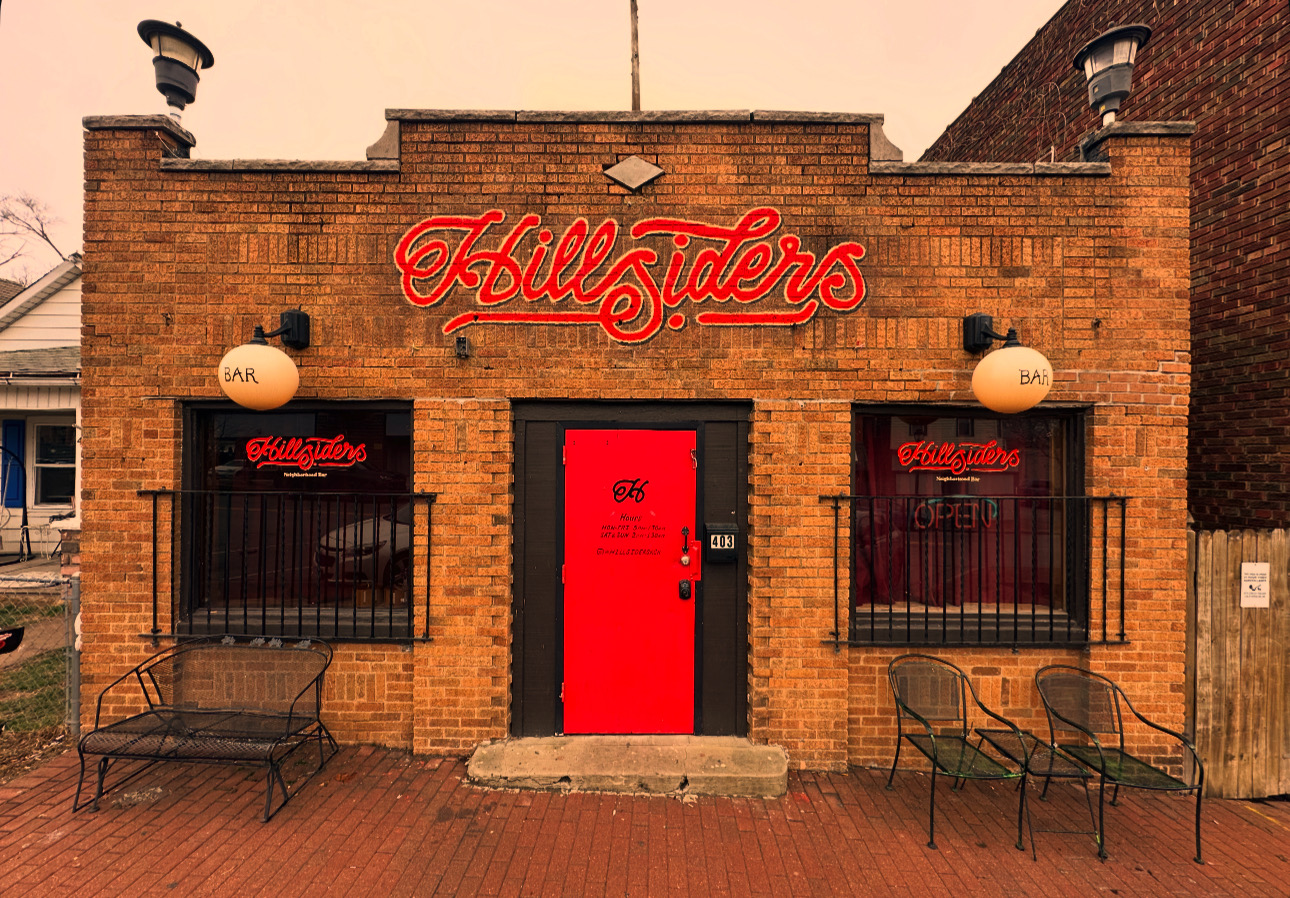 Brick building with red door, neon sign, two windows with black railings, and outdoor seating.