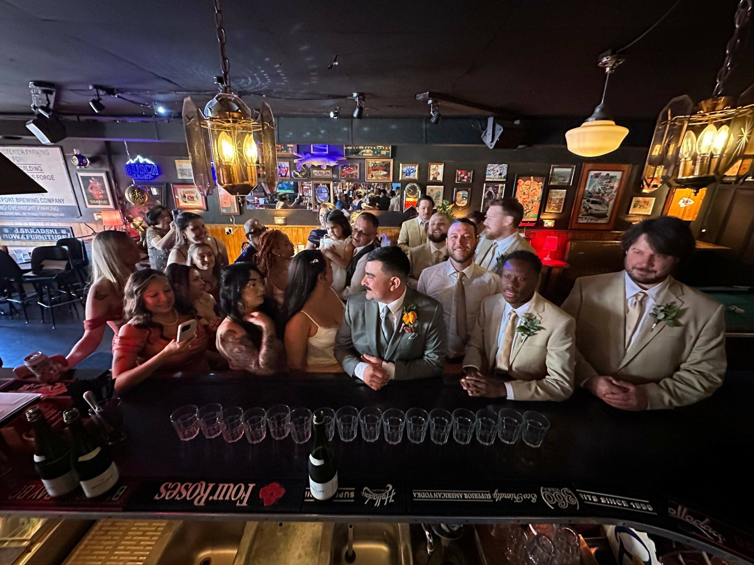 People gathered in a bar, some dressed formally, with a DJ and bar counter in the background.
