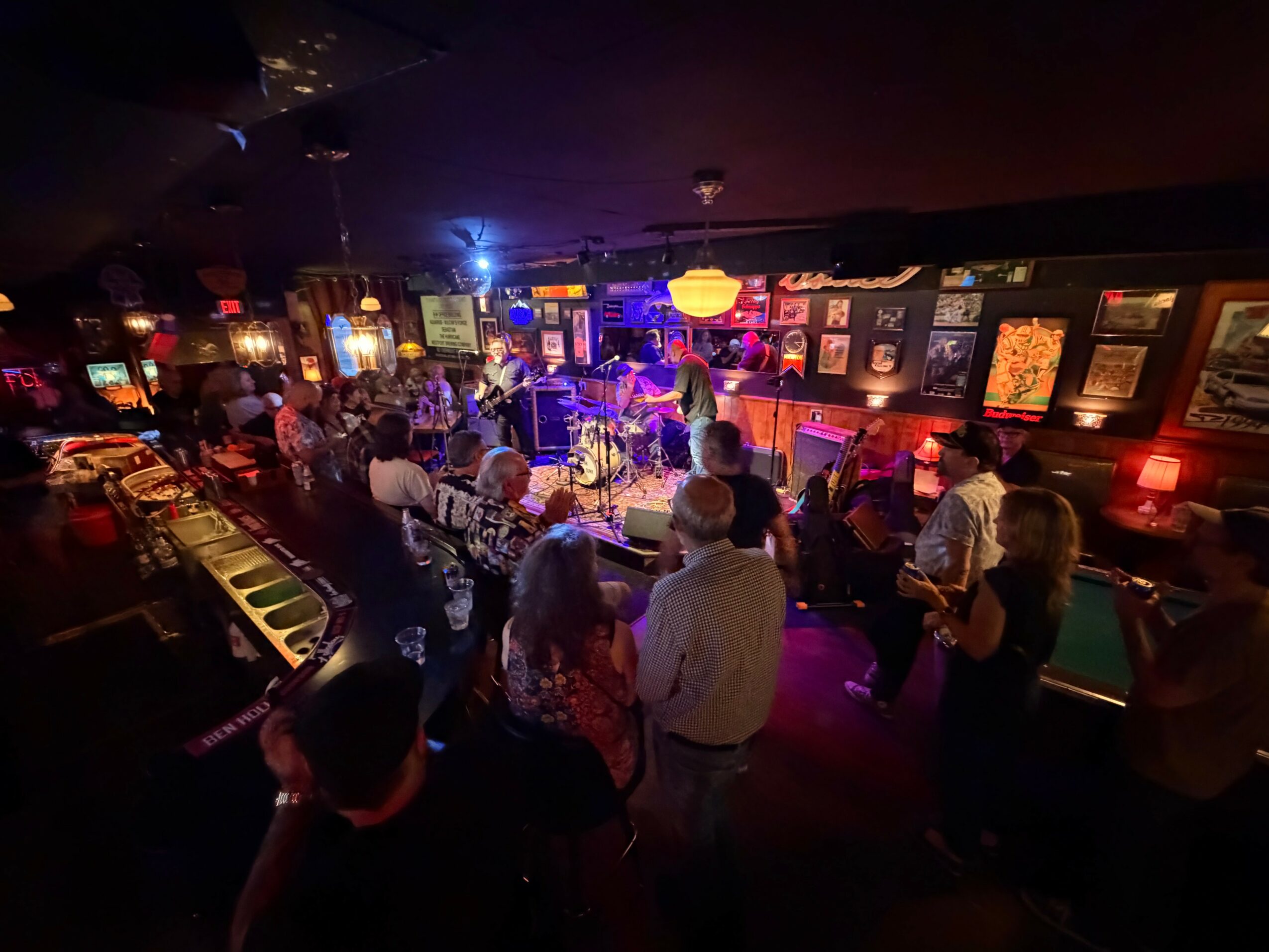 People gather in a dimly lit bar with a stage, colorful lights, and artwork on the walls, enjoying live music.