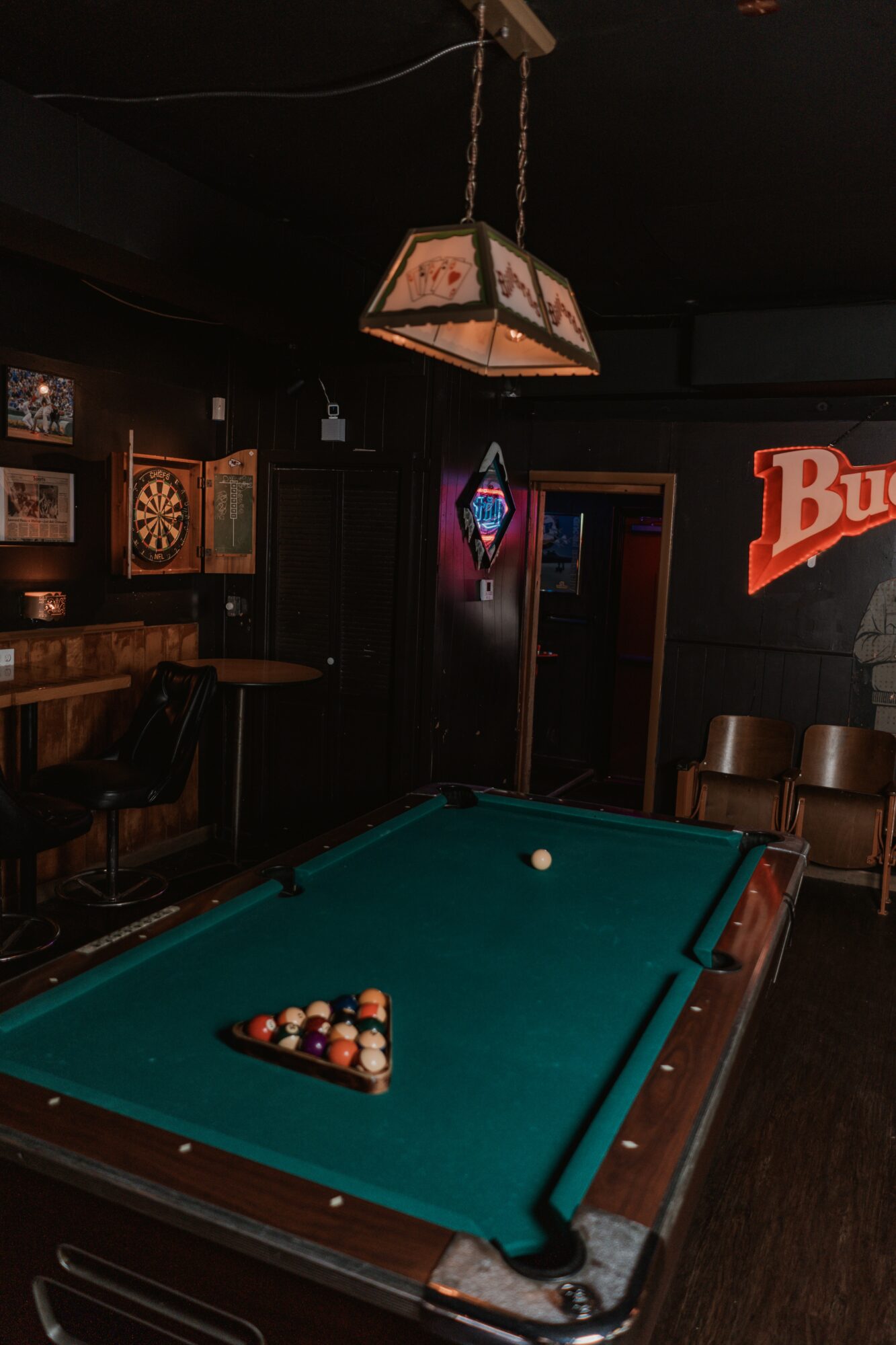Pool table with balls racked, hanging light fixture, dartboard, and bar area in a dimly lit pub setting.