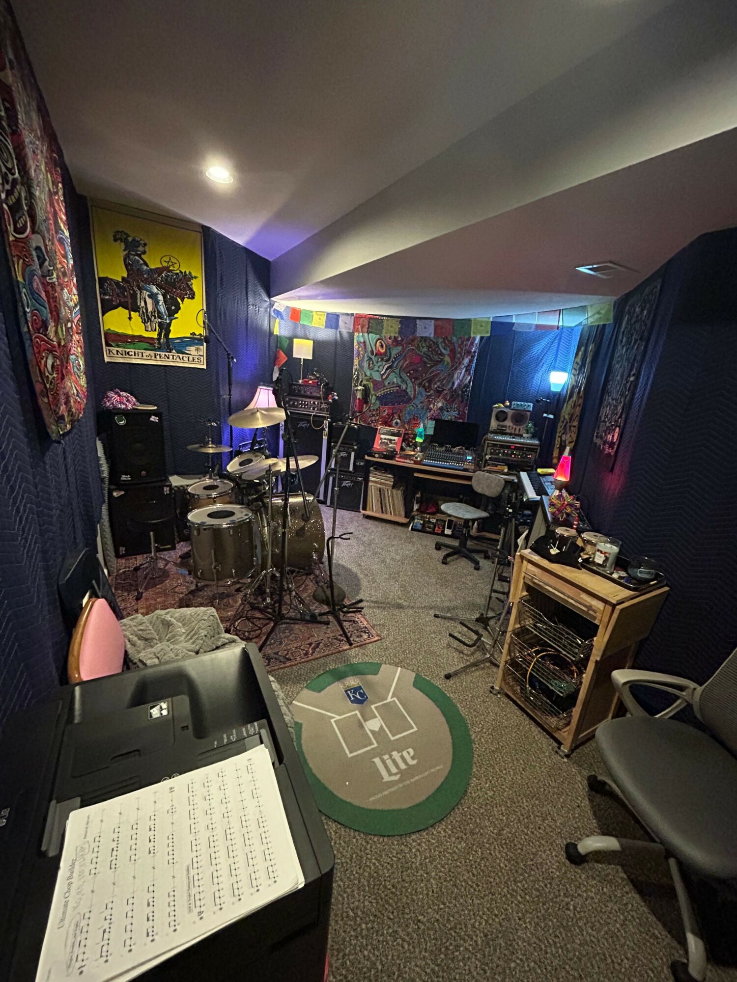 Music studio with equipment, a chair, and colorful artwork on the wall, viewed from a tilted angle.