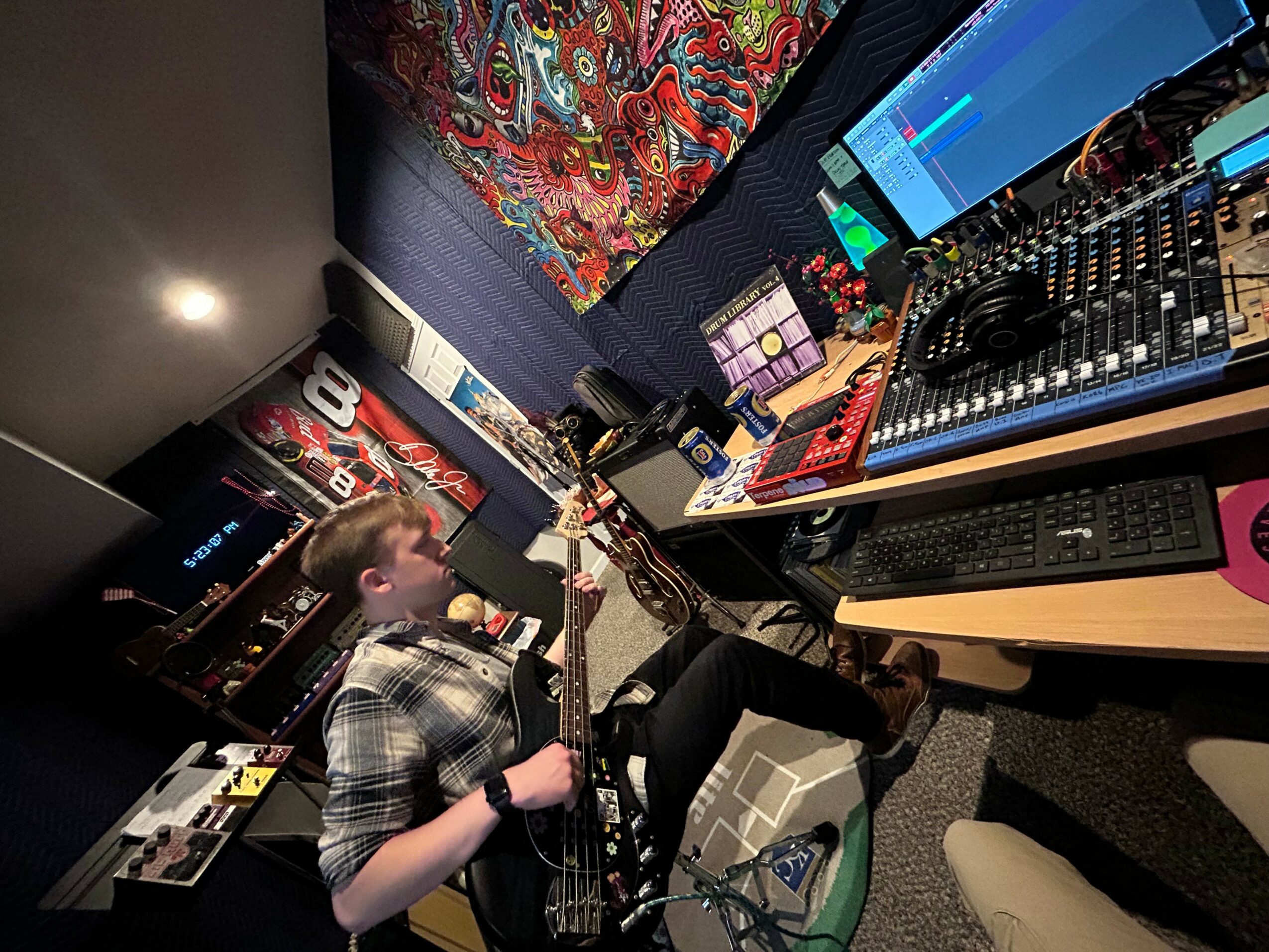 Person working with DJ equipment and computer screens in a music studio, colorful fabric in background.