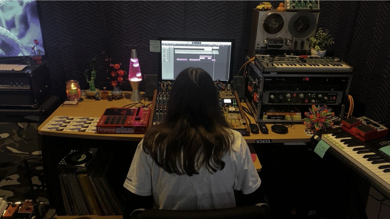 Person with long hair working at a music production setup with a computer, keyboard, and various electronic equipment in a studio.