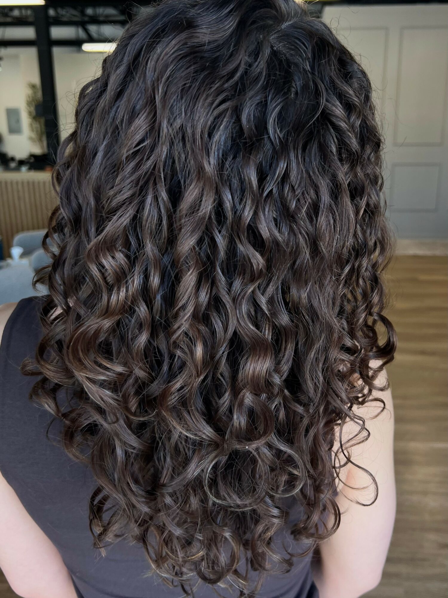 Person with long, dark, curly hair viewed from the back, indoors, with a wooden floor and light-colored wall.