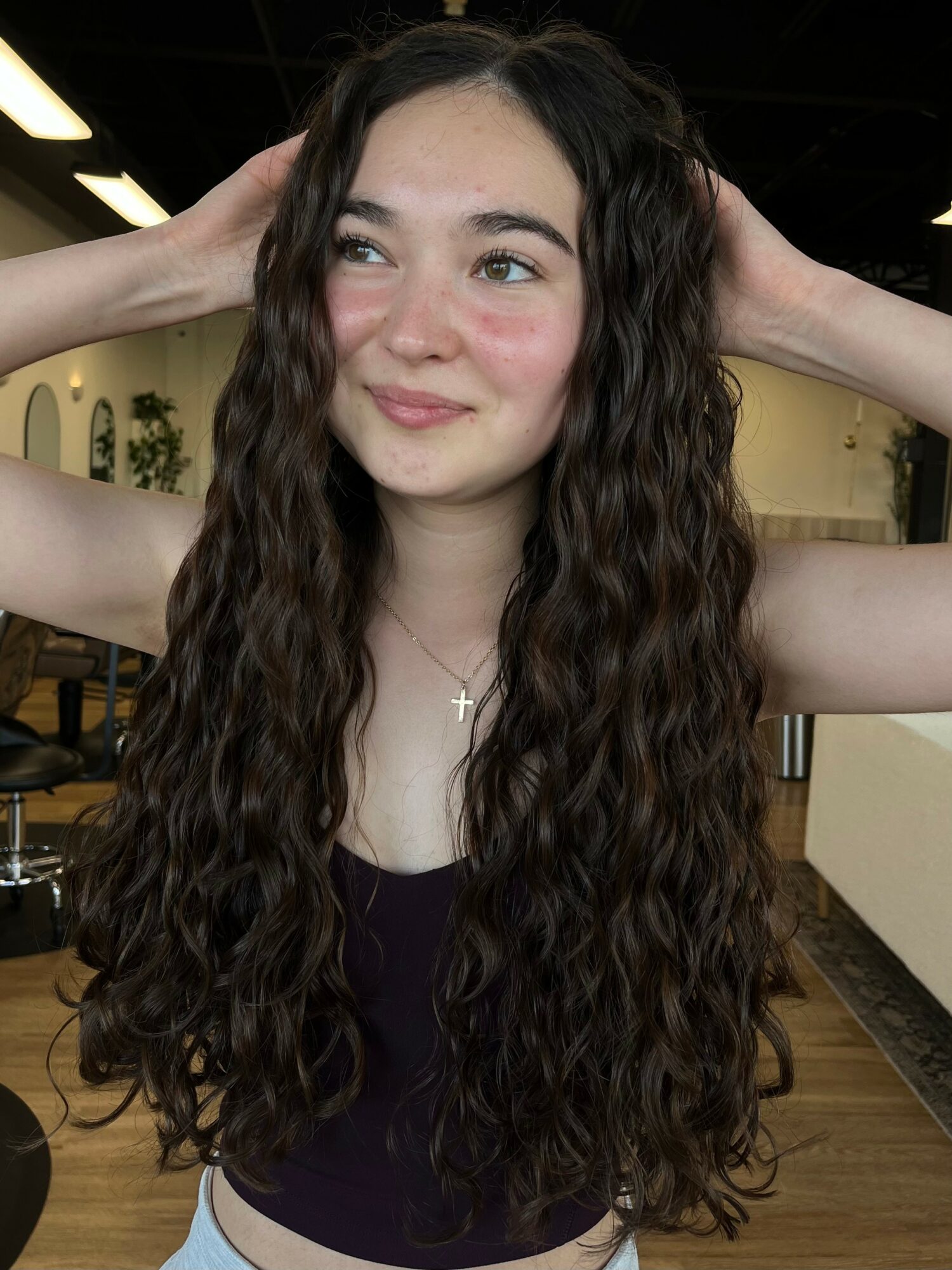 Young woman with long, curly hair smiling, hands behind her head, wearing a necklace with a cross, indoors.