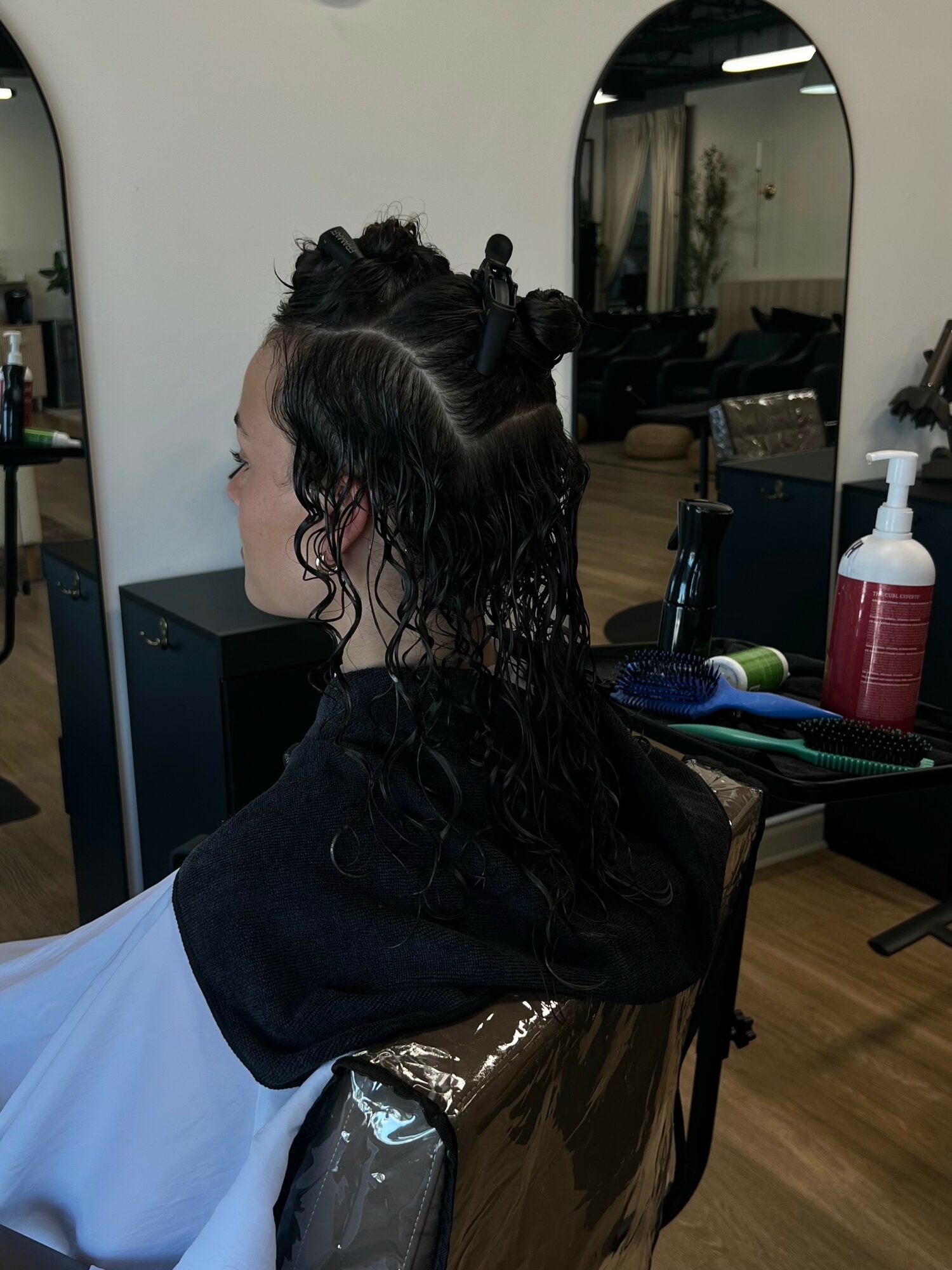 Person with wet, curly hair sitting in a salon chair, facing left, with mirrors and salon tools in background.