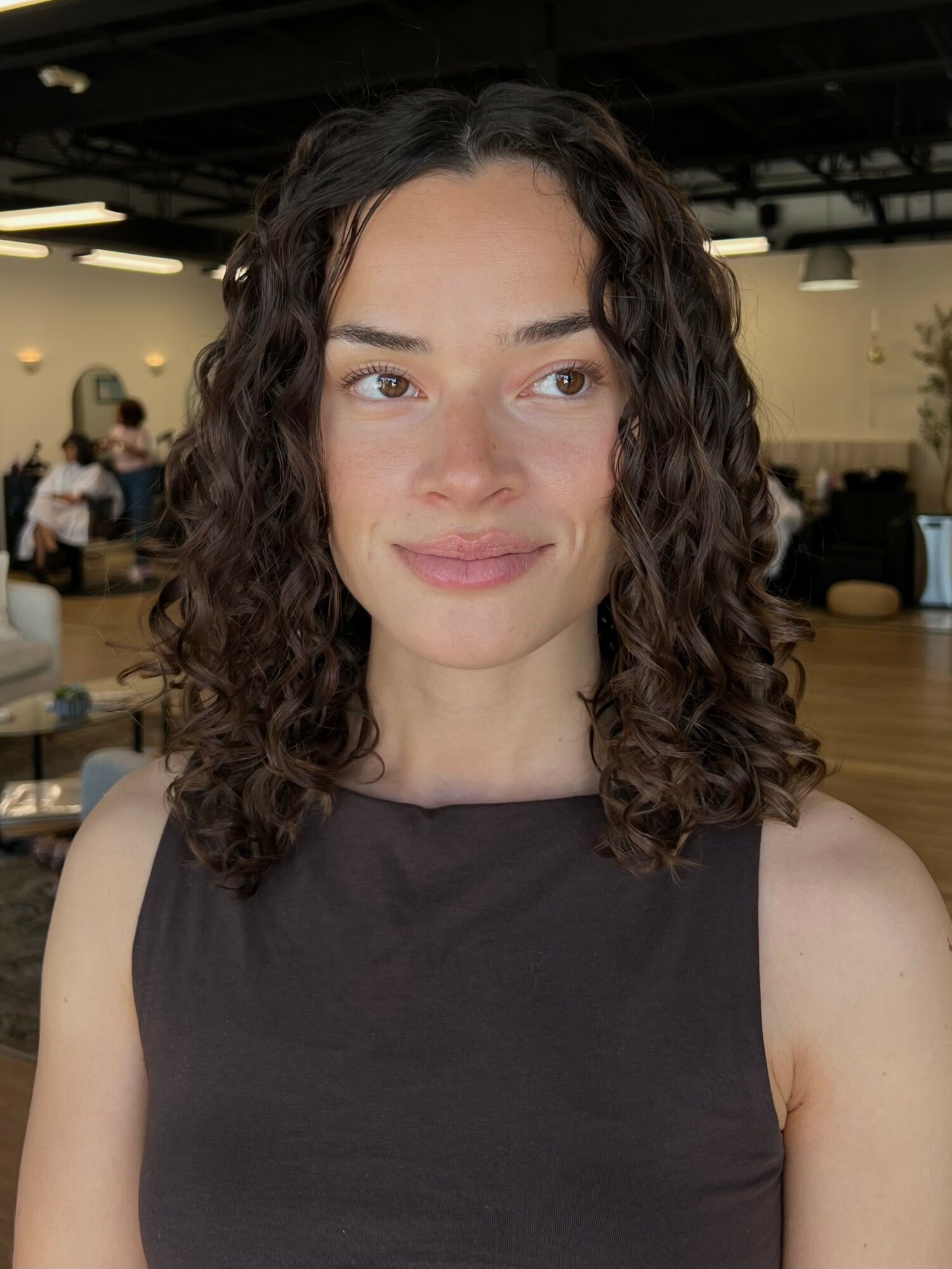 Young woman with curly hair and a sleeveless black top, smiling softly indoors.