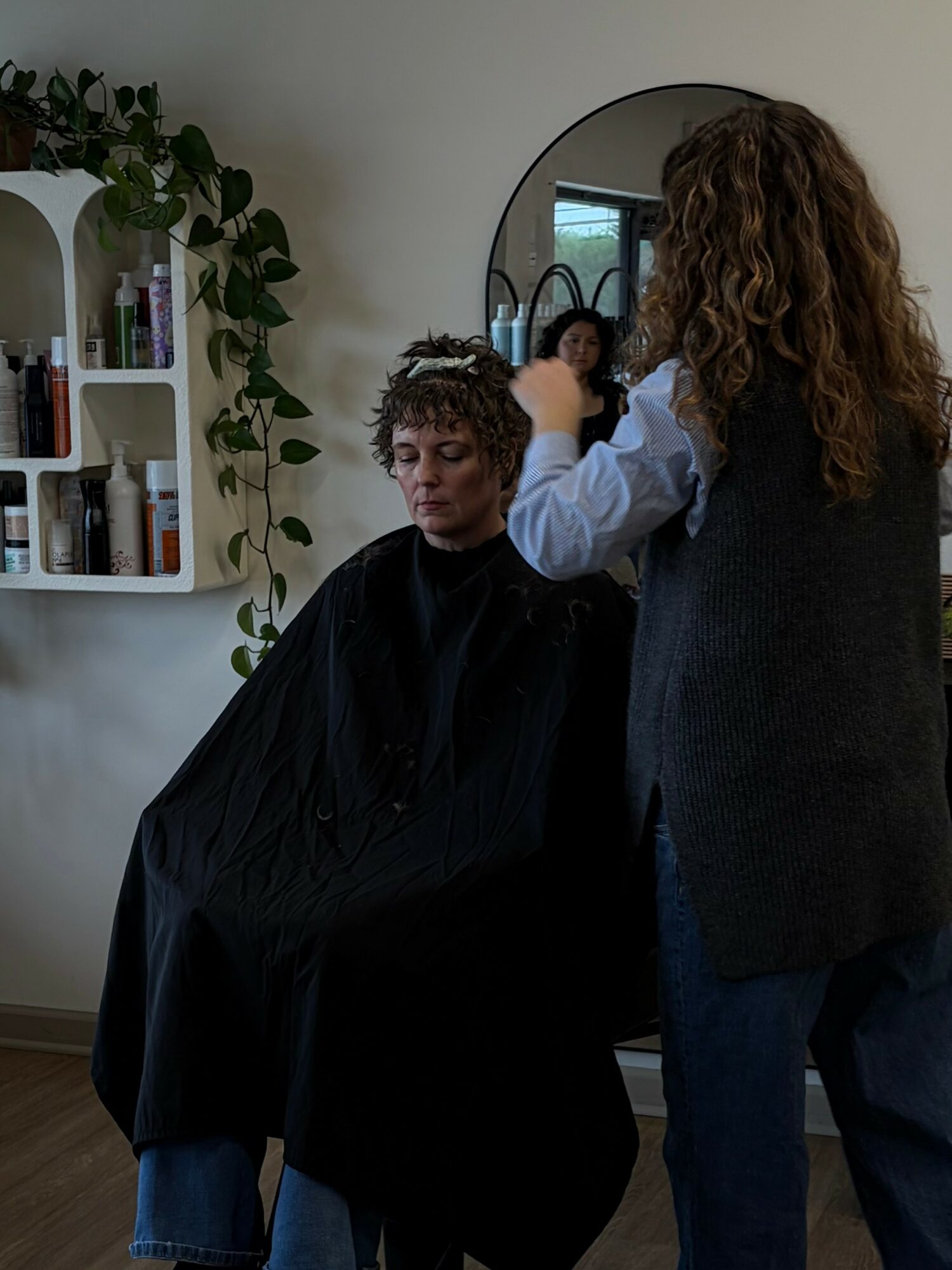 Person getting a haircut in a salon, with a mirror reflecting another person, and shelves with hair products nearby.