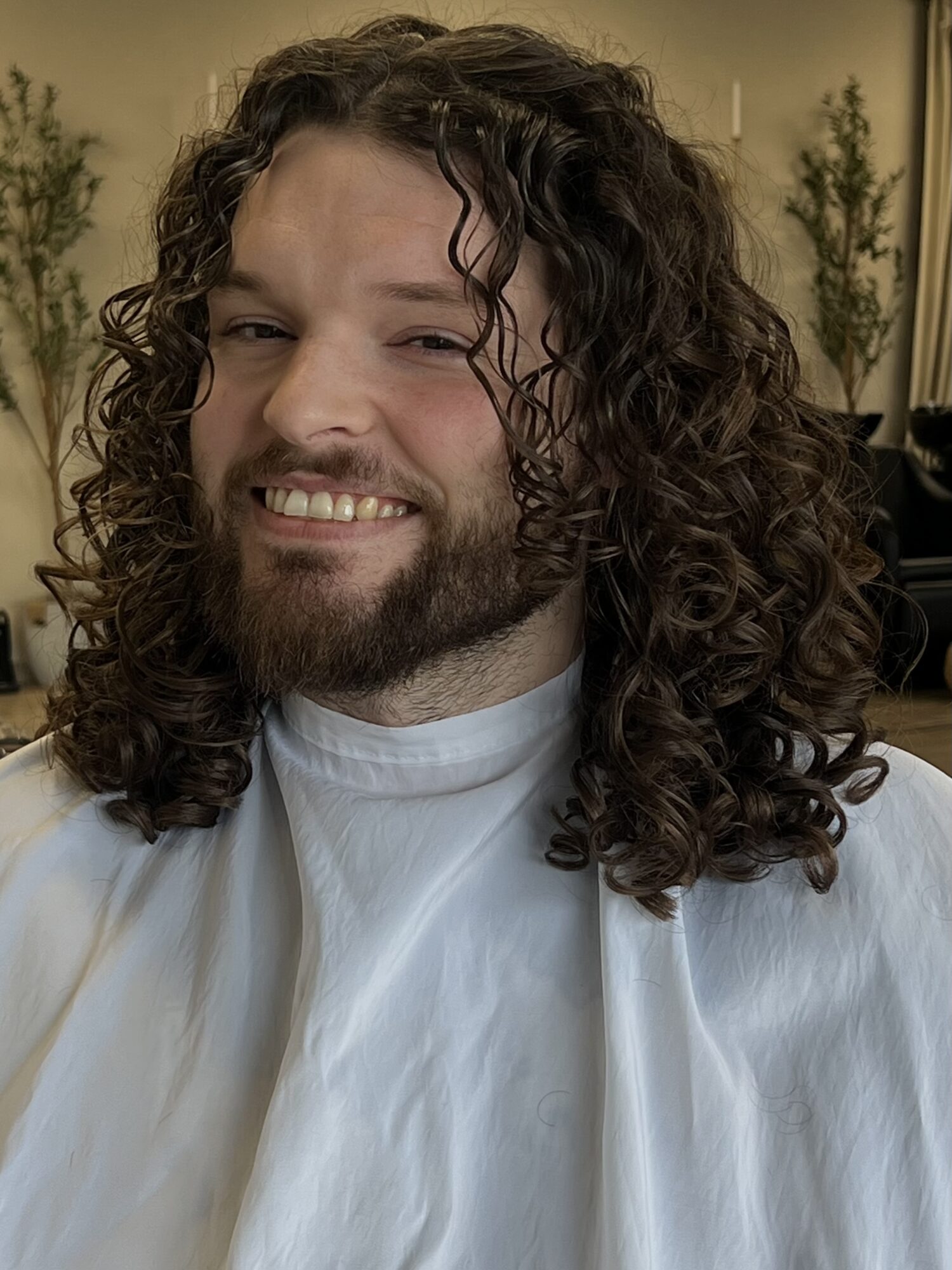 Person with long curly hair and beard smiling, wearing a white cape, in an indoor setting with plants in the background.