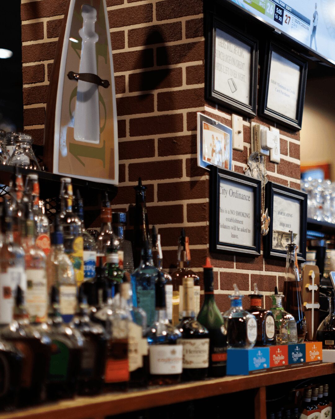 Bar with bottles and framed menus on brick wall, illuminated by overhead lights.
