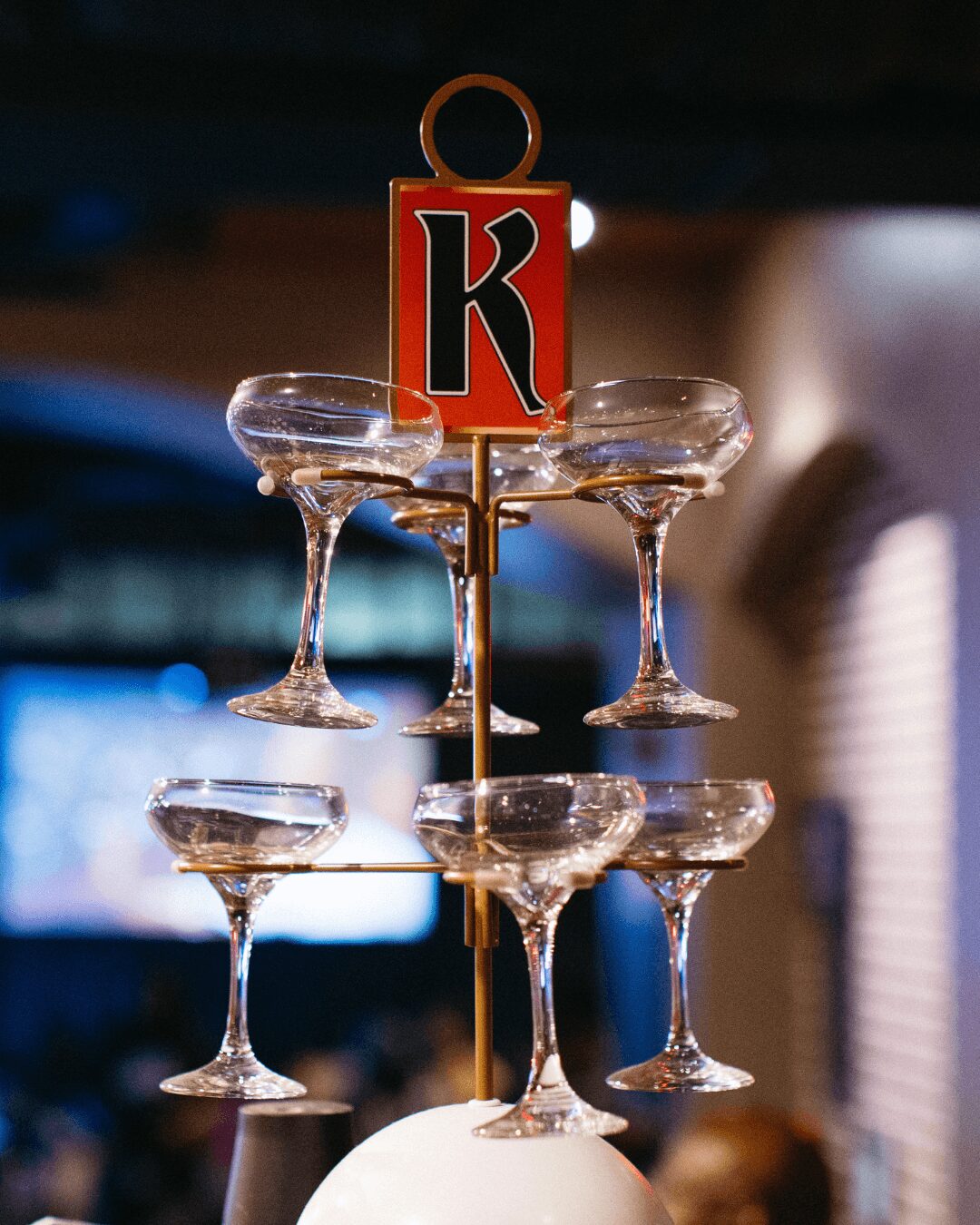 A tower of six empty cocktail glasses with a red and black 'K' sign on top, set against a dark background.