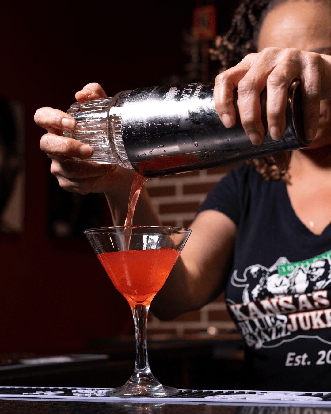 Person pours red cocktail from shaker into a martini glass, with a brick wall background.