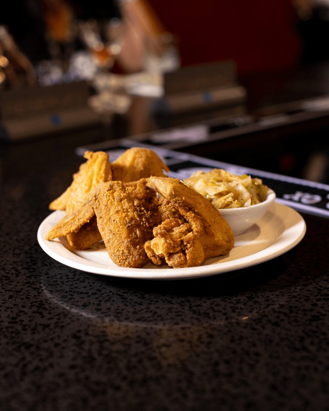 Plate with fried chicken pieces and a side of coleslaw on a dark countertop, background blurred.