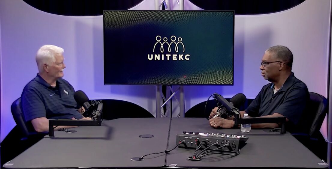 Two men sit at a table with microphones, facing each other, in a studio with a UNITEKC logo on a screen behind them.