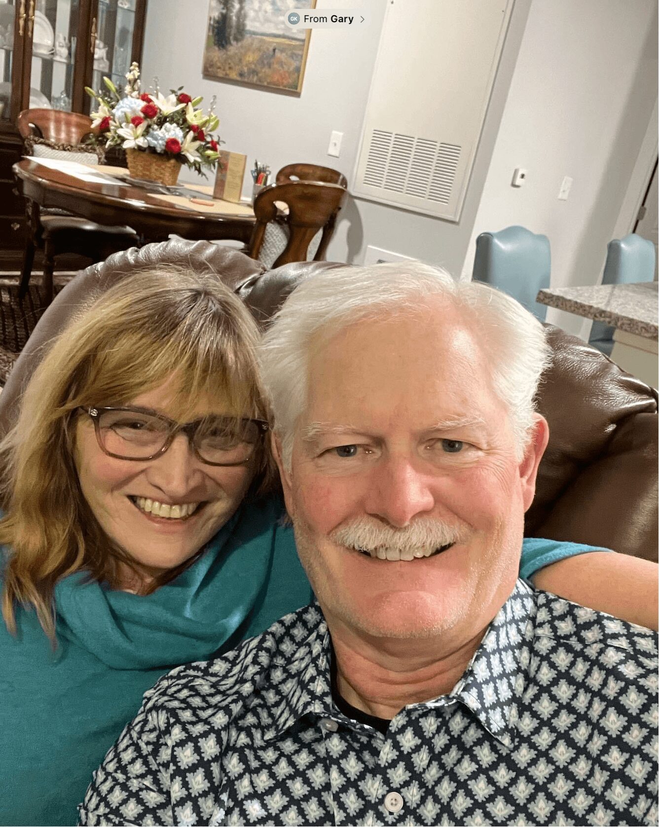 Two smiling people, a woman with glasses and a man with white hair, sitting close together indoors.