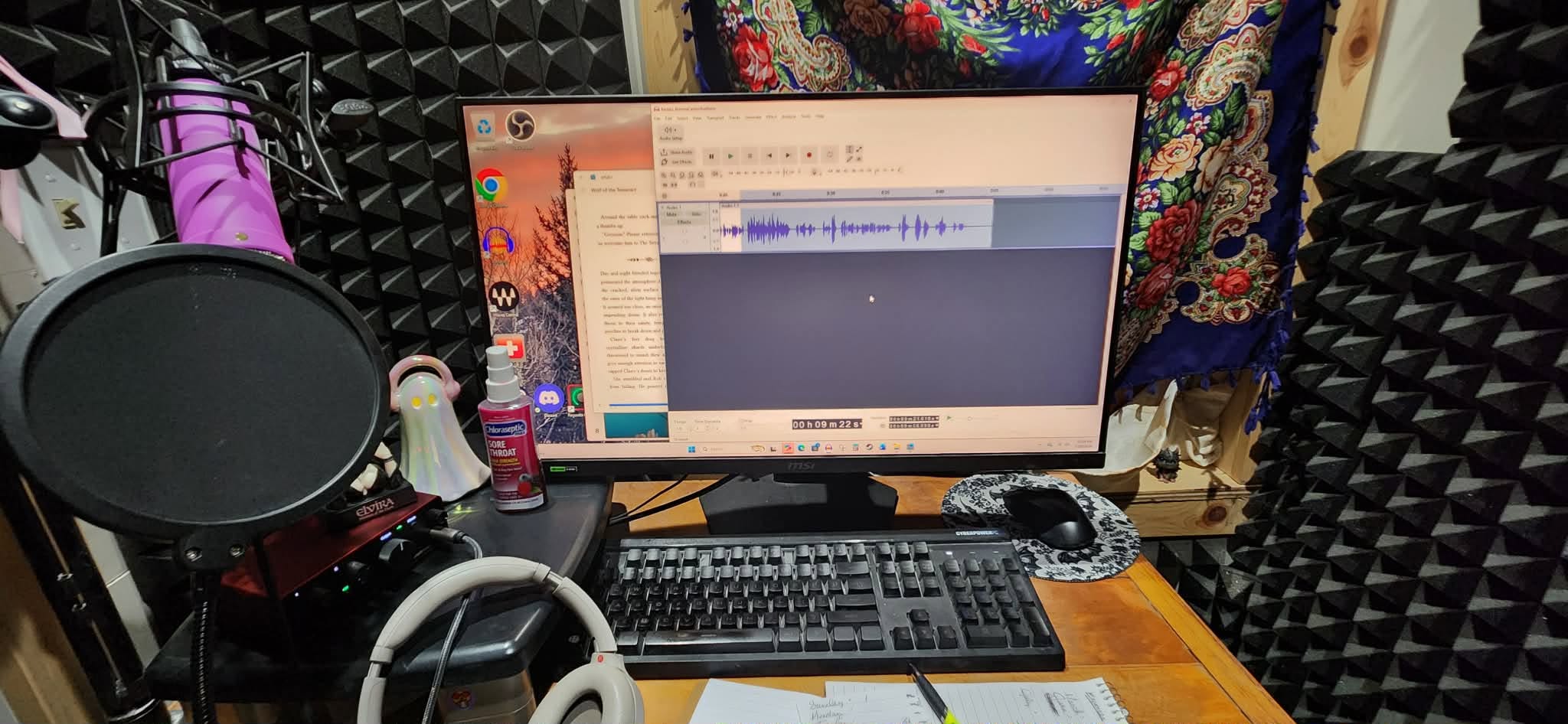 Computer monitor displaying audio editing software, microphone, keyboard, and headphones on a desk, with soundproofing foam on walls.
