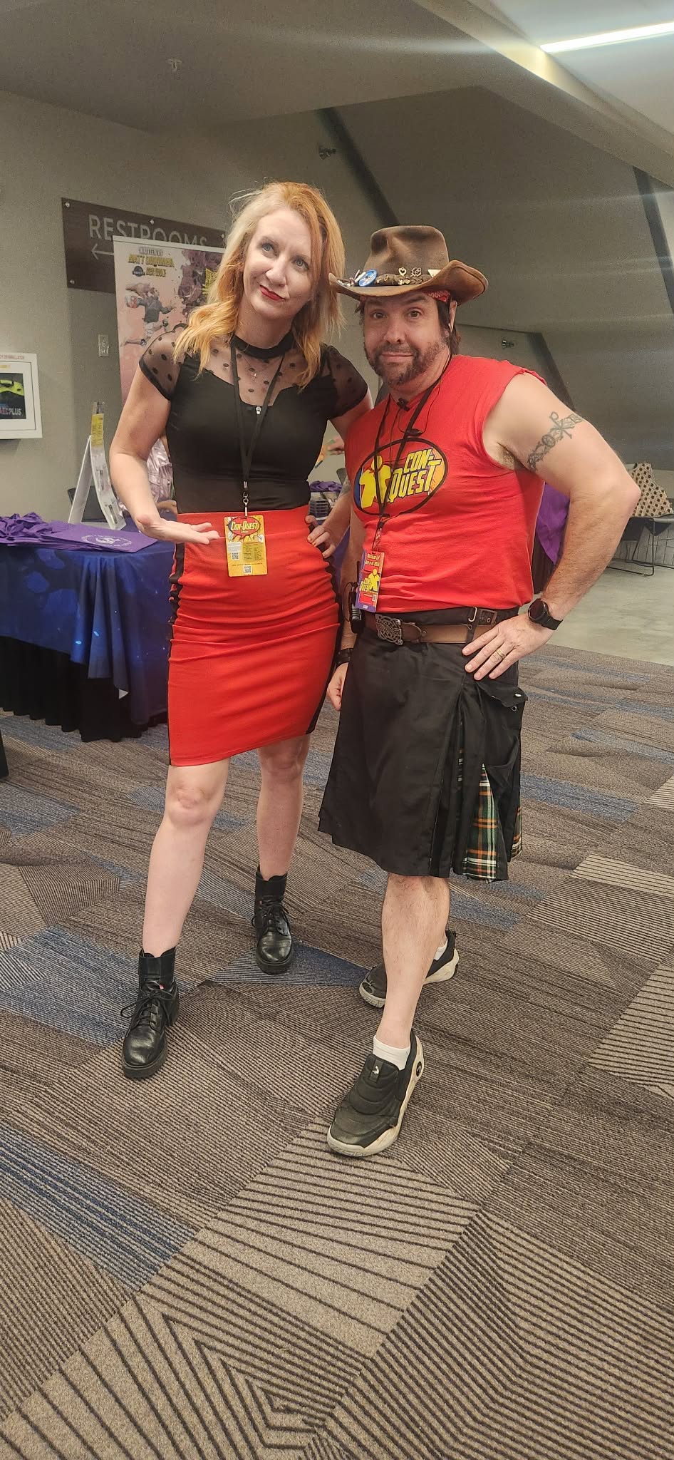 Two people standing together, smiling, one wearing a red skirt and black top, the other in a red shirt, shorts, and a hat.