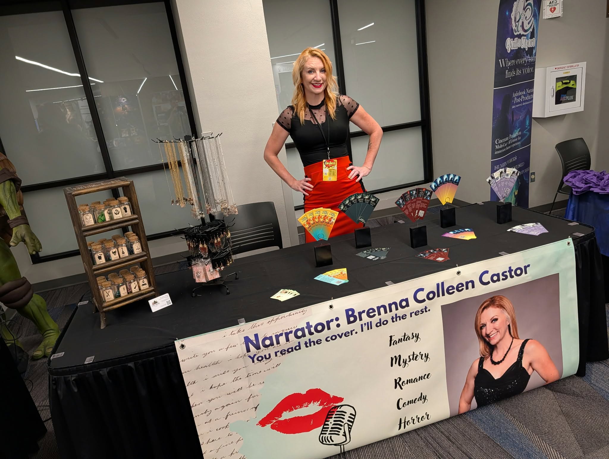 Woman standing behind a table with a banner, in an indoor setting, smiling with hands on hips, books and decorations on table.