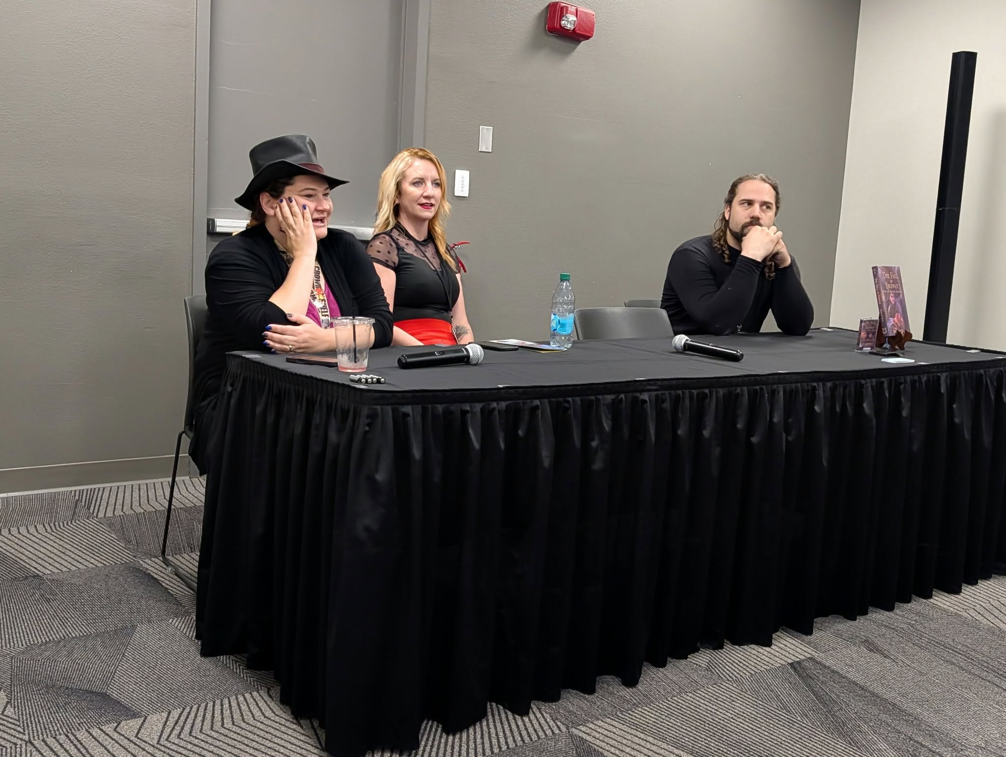 Three people sitting at a long table with black tablecloth in a conference room, two women and one man, with water bottles and electronic devices.