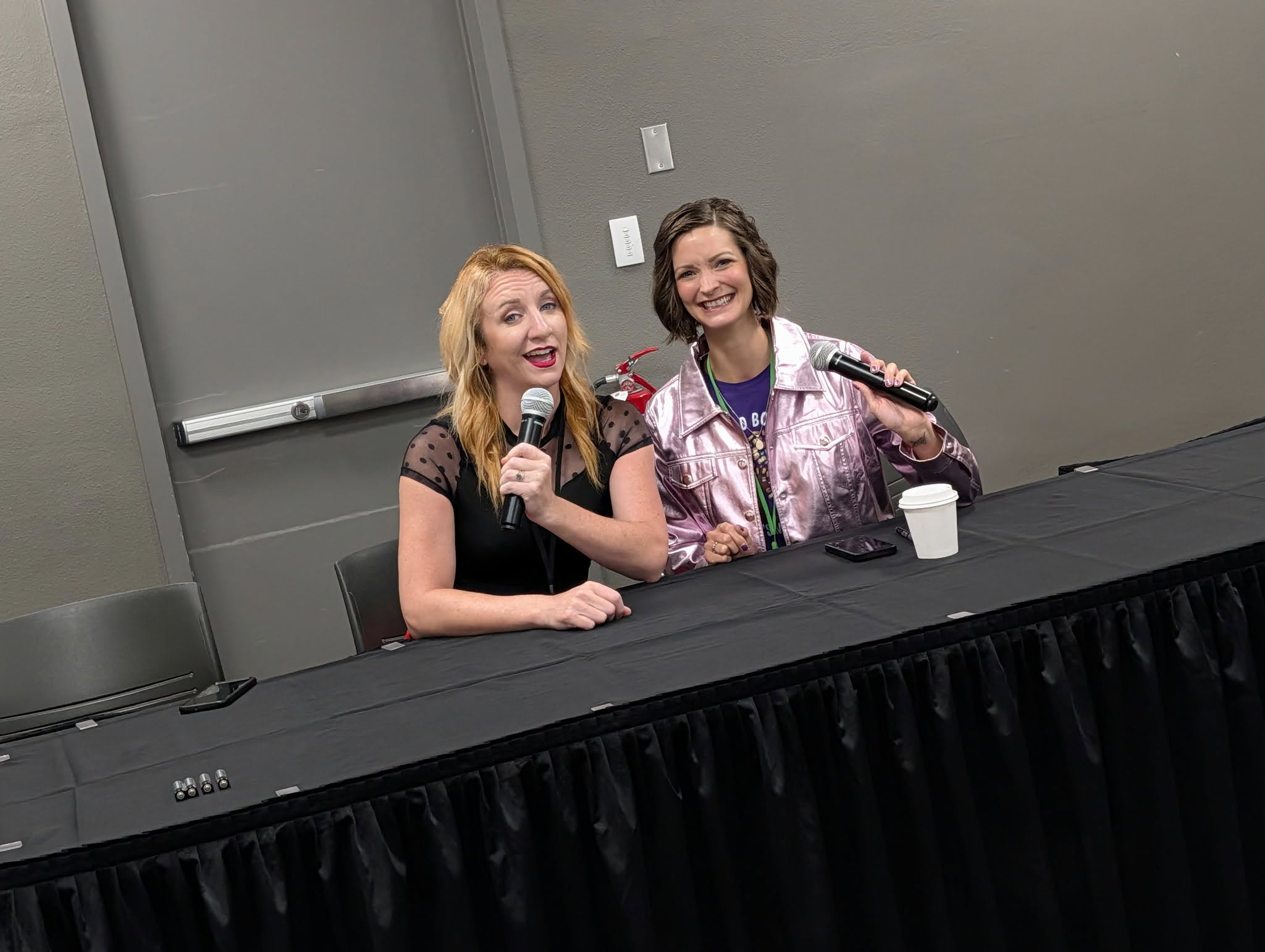 Two women sit at a table with microphones, smiling and talking, in a room with a gray wall and a whiteboard.