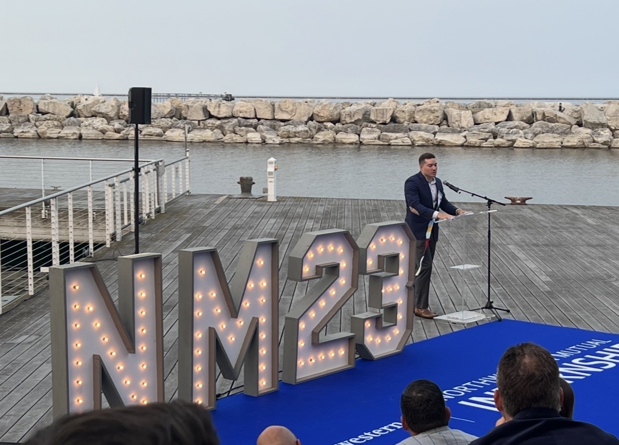 Man in suit speaking at a podium outdoors near water with large illuminated letters NM23, audience in foreground.