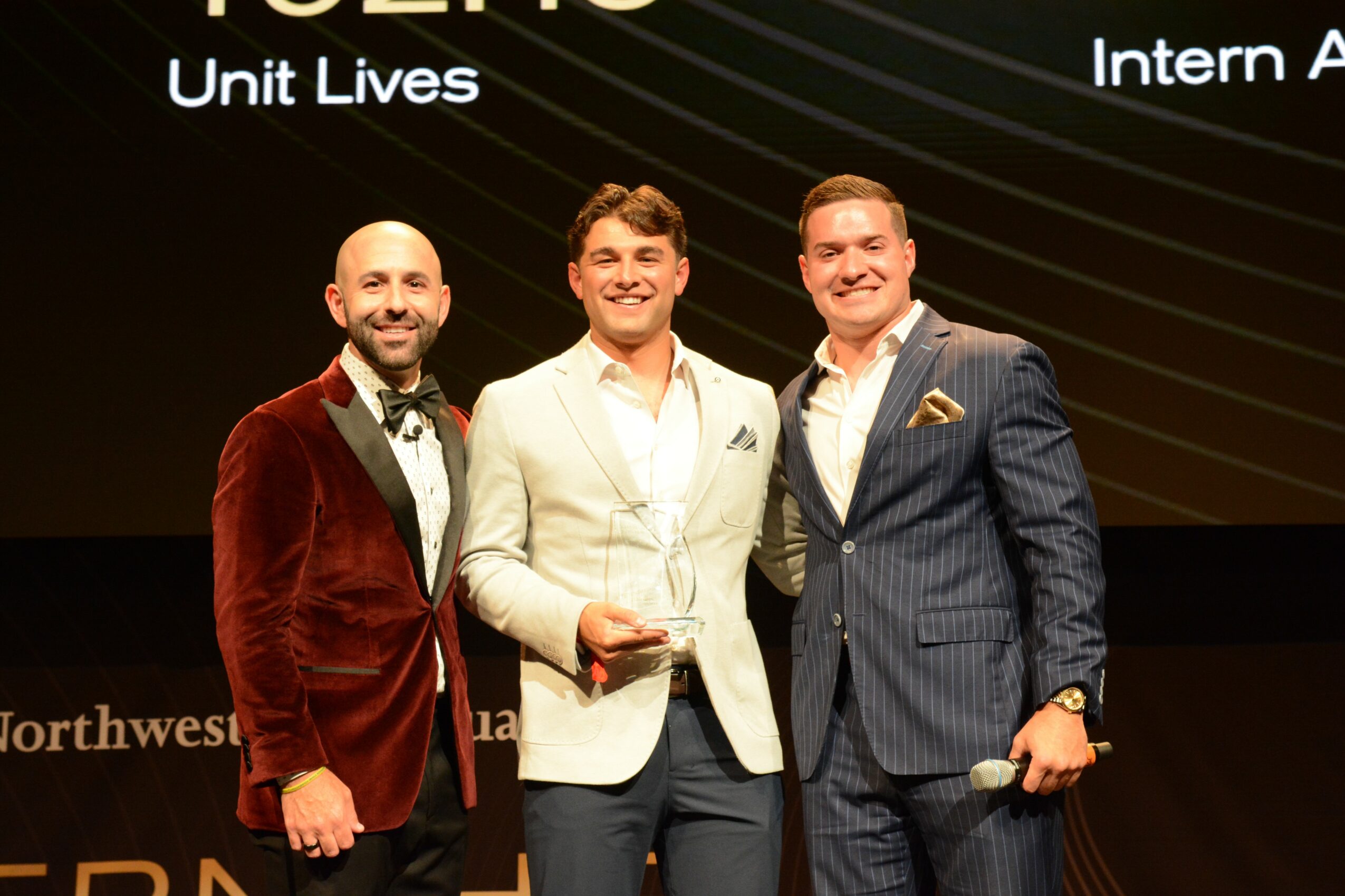 Three men in formal attire standing on stage, smiling, with a dark background and text. One holds an award.