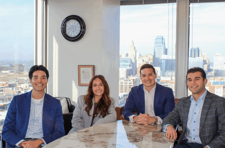 Four people sitting at a table in a modern office with large windows and city view.
