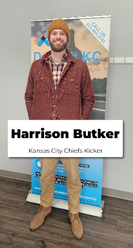 Man with beard wearing a brown beanie, plaid shirt, and red jacket standing in front of a Kansas City Chiefs banner.