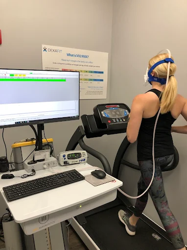 Woman running on treadmill with headphones in a clinical setting, computer monitor and equipment nearby.