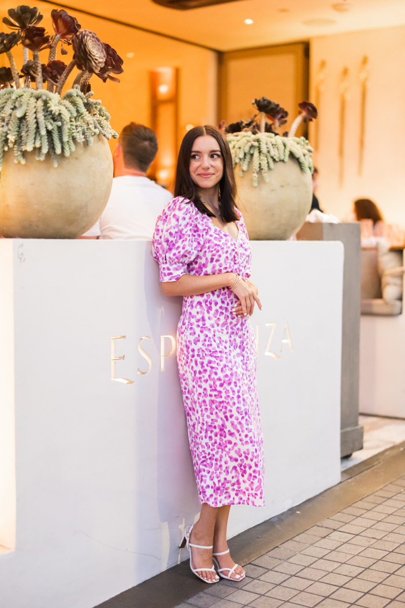 Woman in pink and white patterned dress standing at a reception desk with large flower pots, smiling, in an indoor setting.