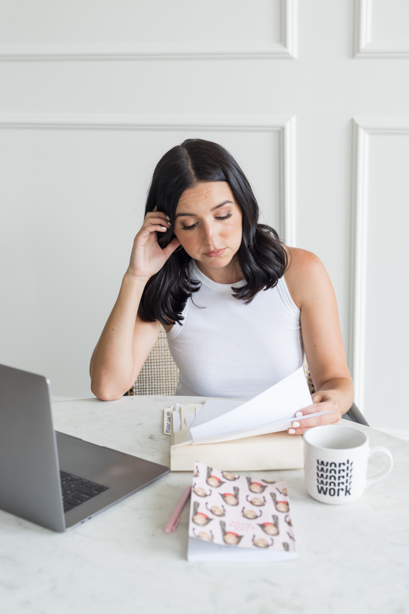 Woman with dark hair reading papers at desk with laptop, mug, and notebook, in a white room.