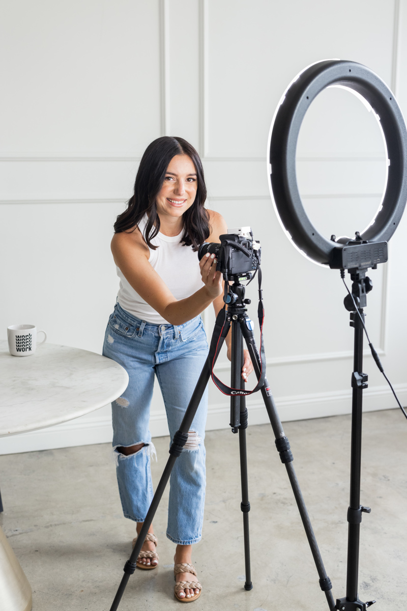 Young woman with dark hair in a white top and jeans stands in a room with a camera and ring light, smiling.