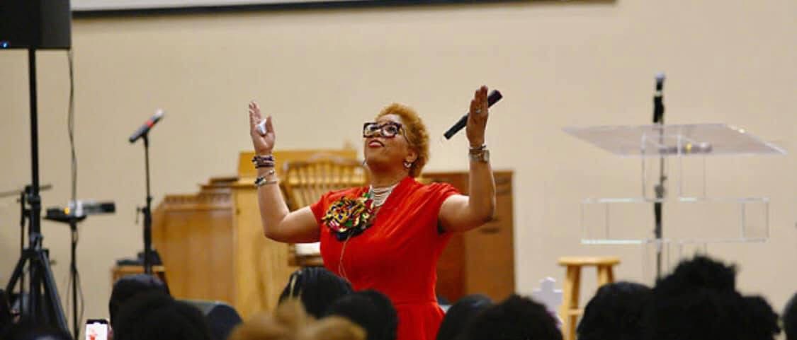 Woman in red dress with glasses conducting or speaking in front of an audience, with musical instruments and microphones in background.