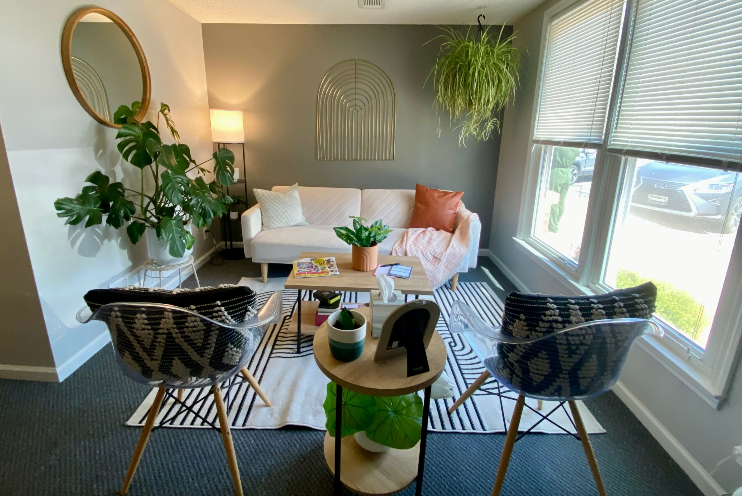 Living room with two chairs, a white sofa, a round mirror, plants, and a window with blinds.