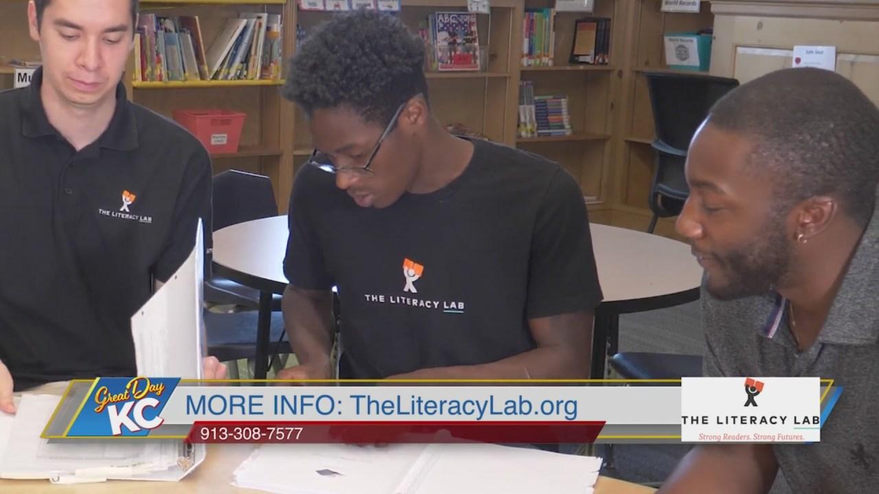 Three people sit at a table in a library, looking at a laptop screen, with bookshelves in the background.