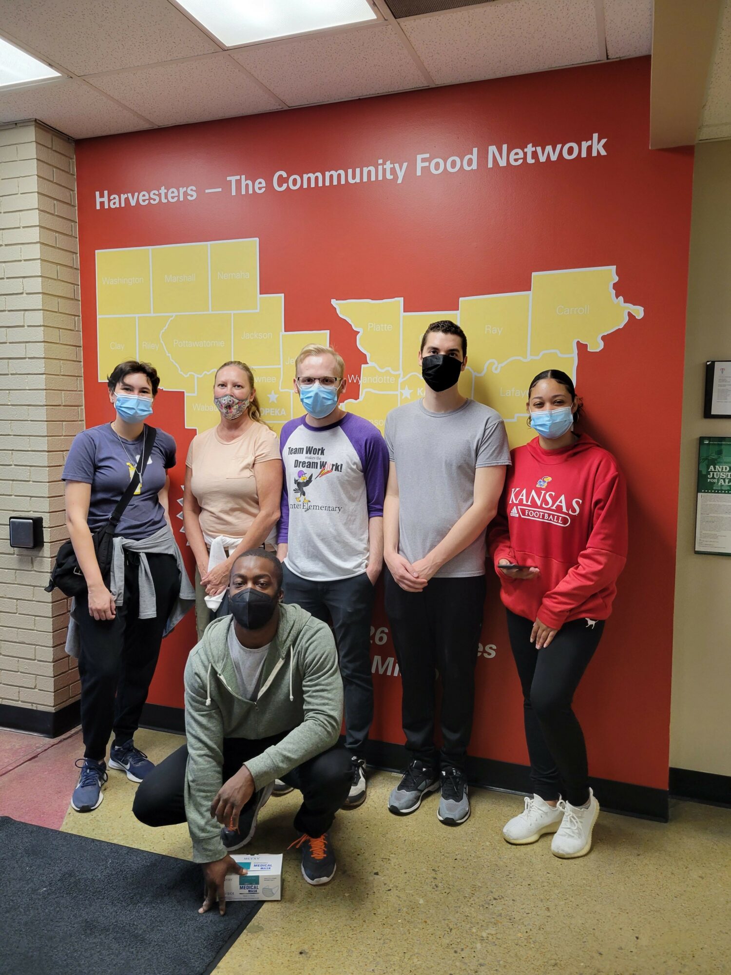Five people standing in front of a red and yellow banner with text, some wearing masks, in an indoor setting.