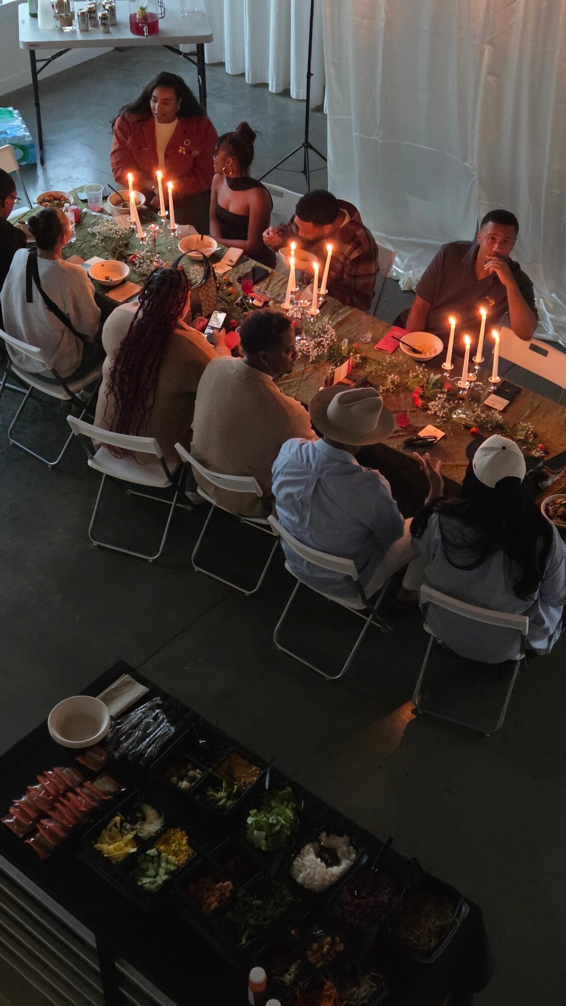 People gathered around a table with candles and food, indoors with a high ceiling and large windows.