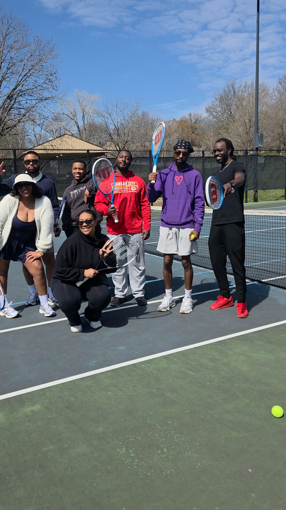 Group of seven people on a tennis court, some holding rackets, with a tennis ball nearby, outdoors on a sunny day.