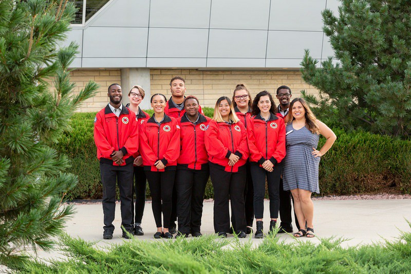 Group of nine people standing outdoors in front of a building, smiling, wearing red jackets, with trees and bushes around.