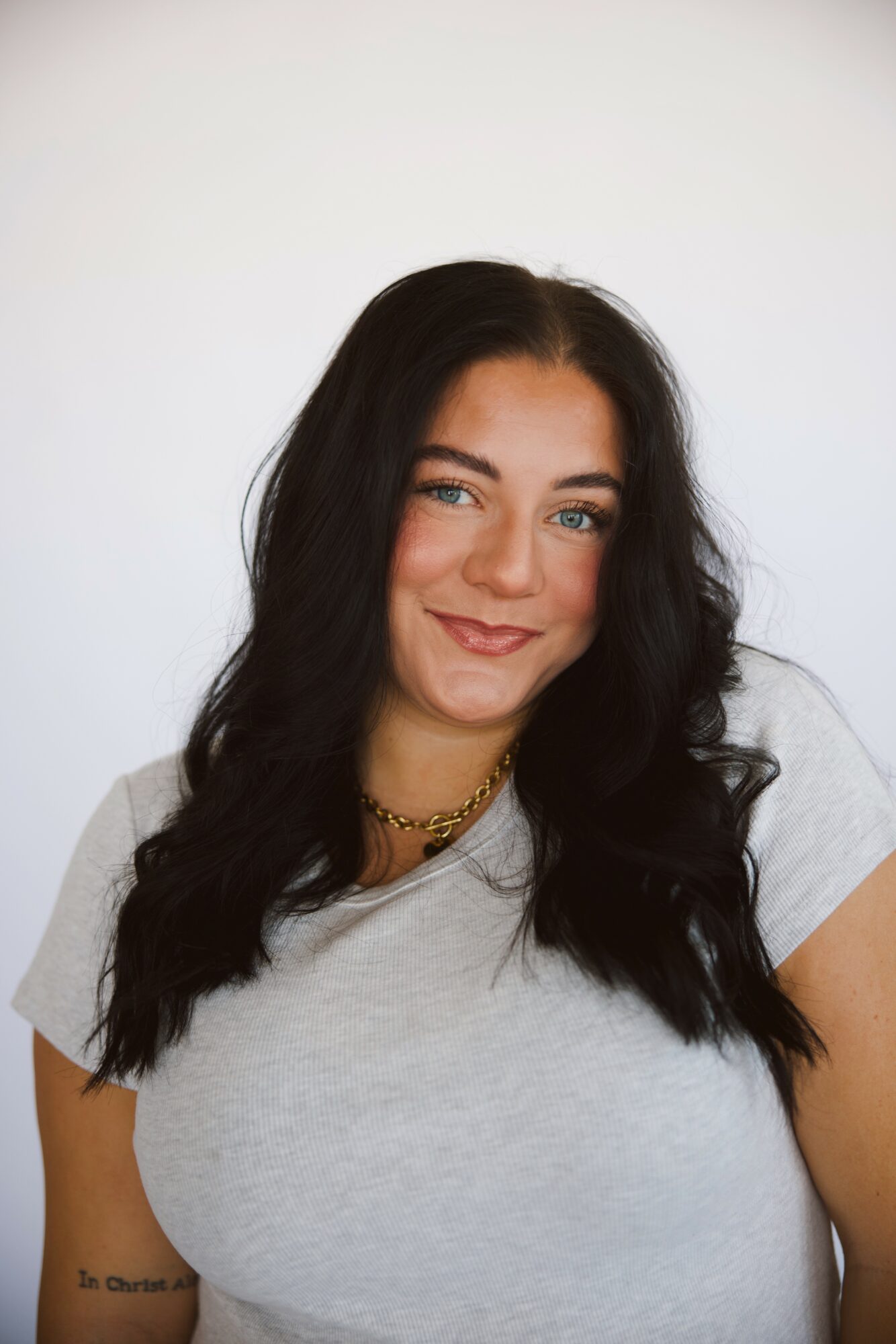 Young woman with long dark hair, smiling, wearing a light gray t-shirt and a gold necklace, against a plain white background.