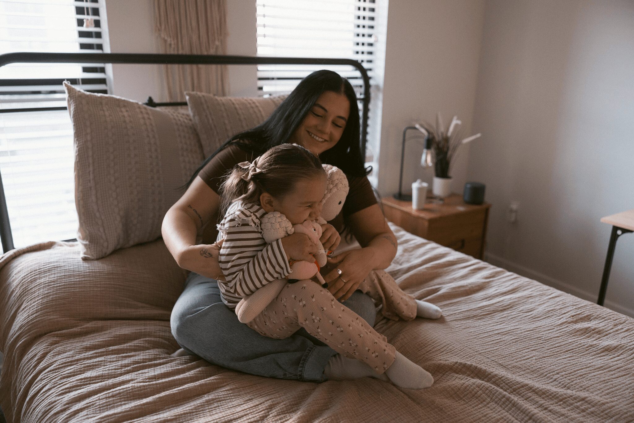 Woman sitting on bed with two children, smiling, in a bedroom with window blinds and plants, daytime.