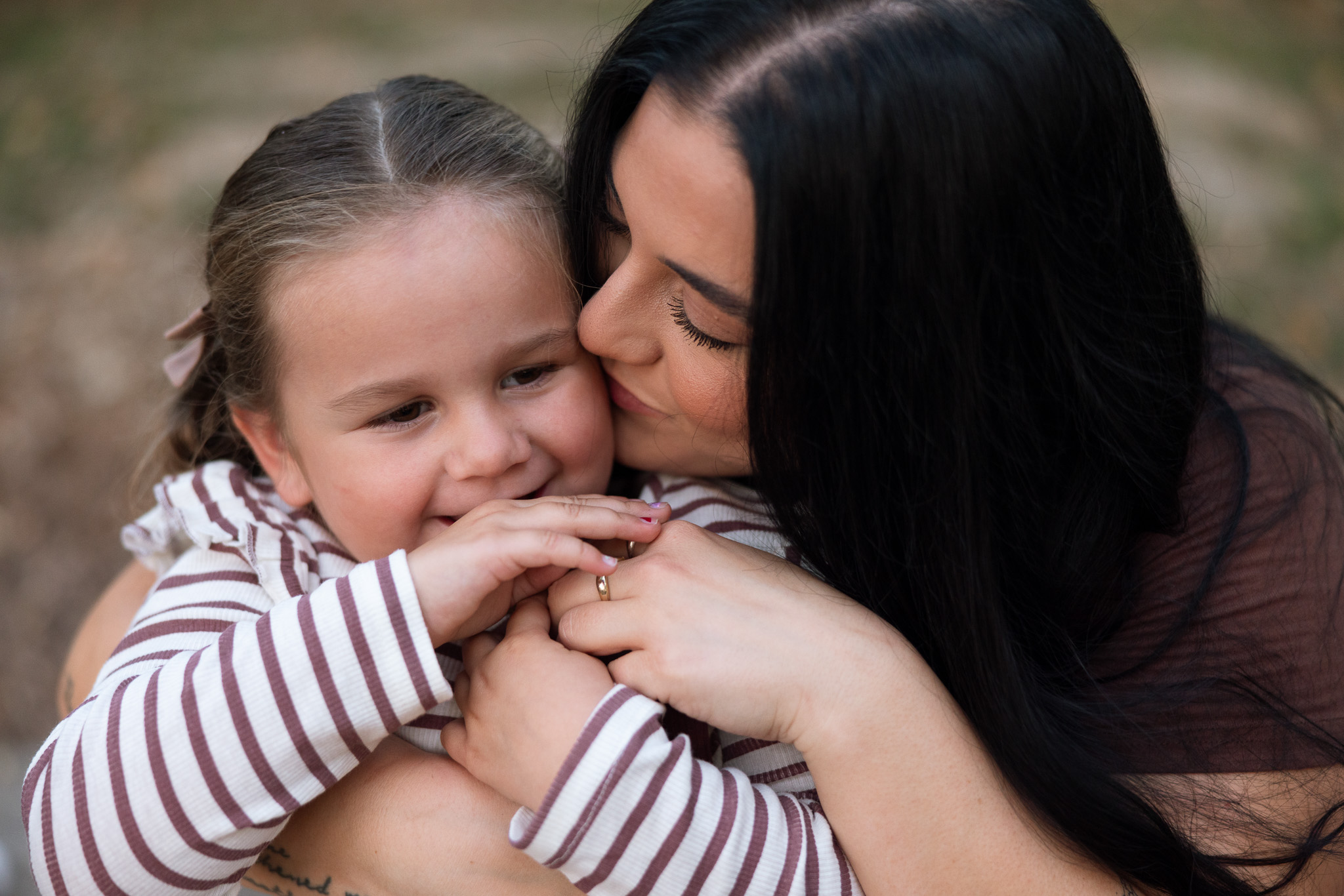 Woman and young girl hugging outdoors, smiling, girl wearing striped shirt, woman with dark hair, affectionate moment.