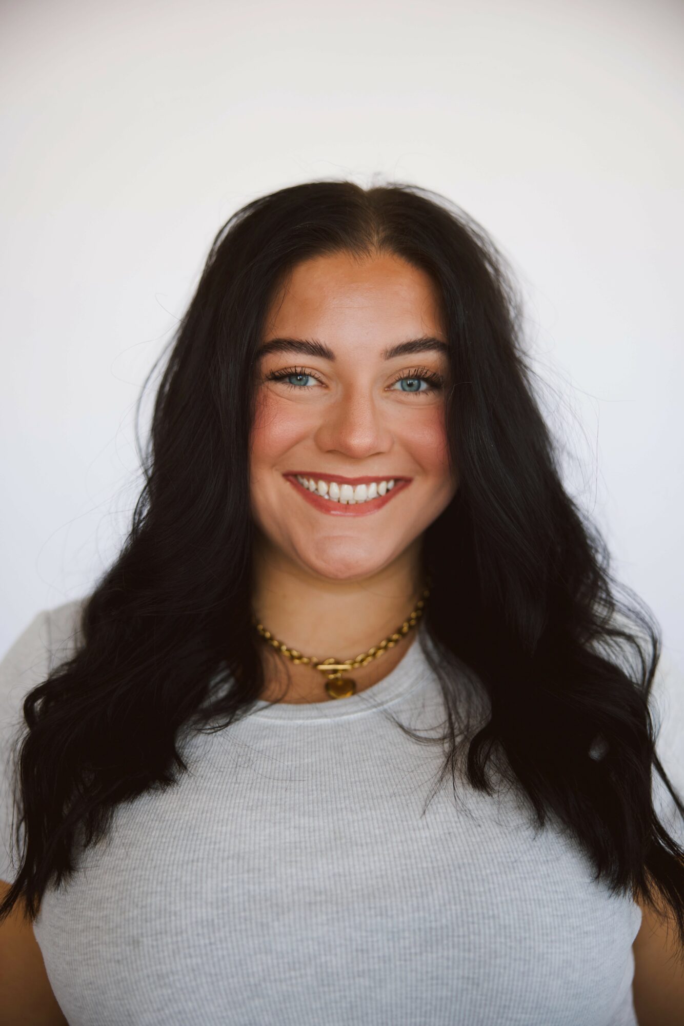 Young woman with long dark hair smiling, wearing a light gray top and a gold necklace, against a plain white background.