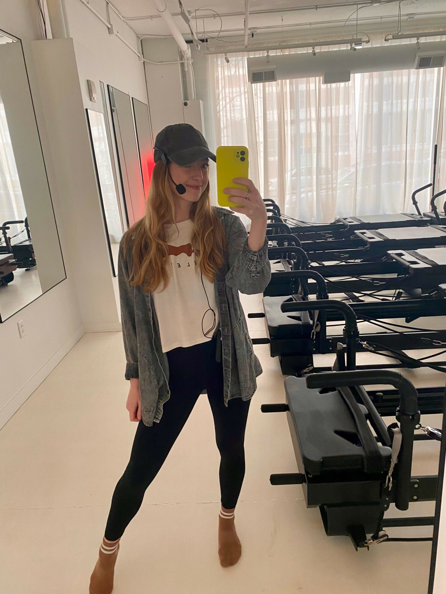 Person taking a mirror selfie in a room with exercise equipment, wearing a cap, glasses, gray cardigan, white top, black leggings, and brown shoes.