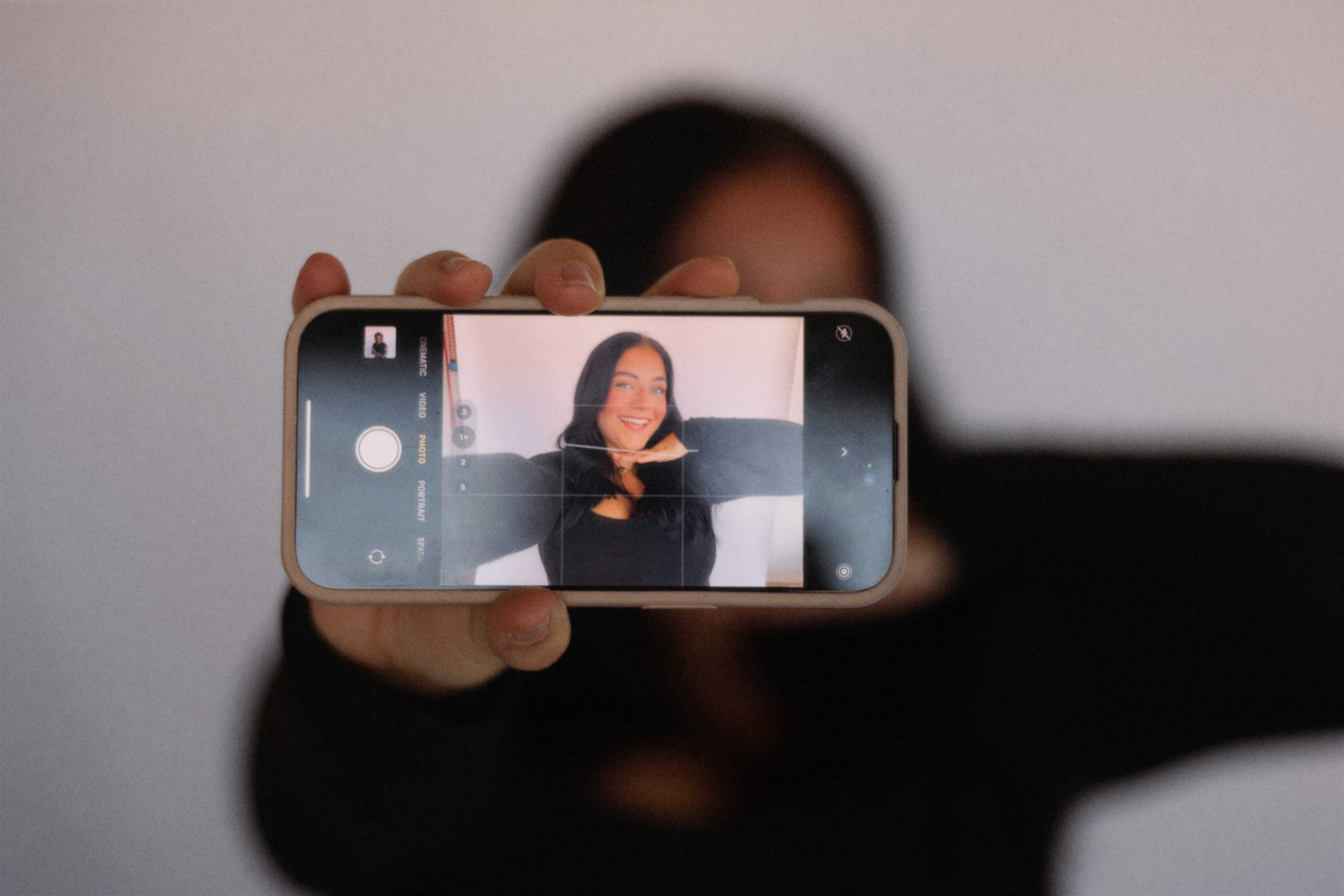 Person holding a smartphone taking a selfie, face partially visible, background plain, phone screen shows the selfie, person wearing dark clothing.