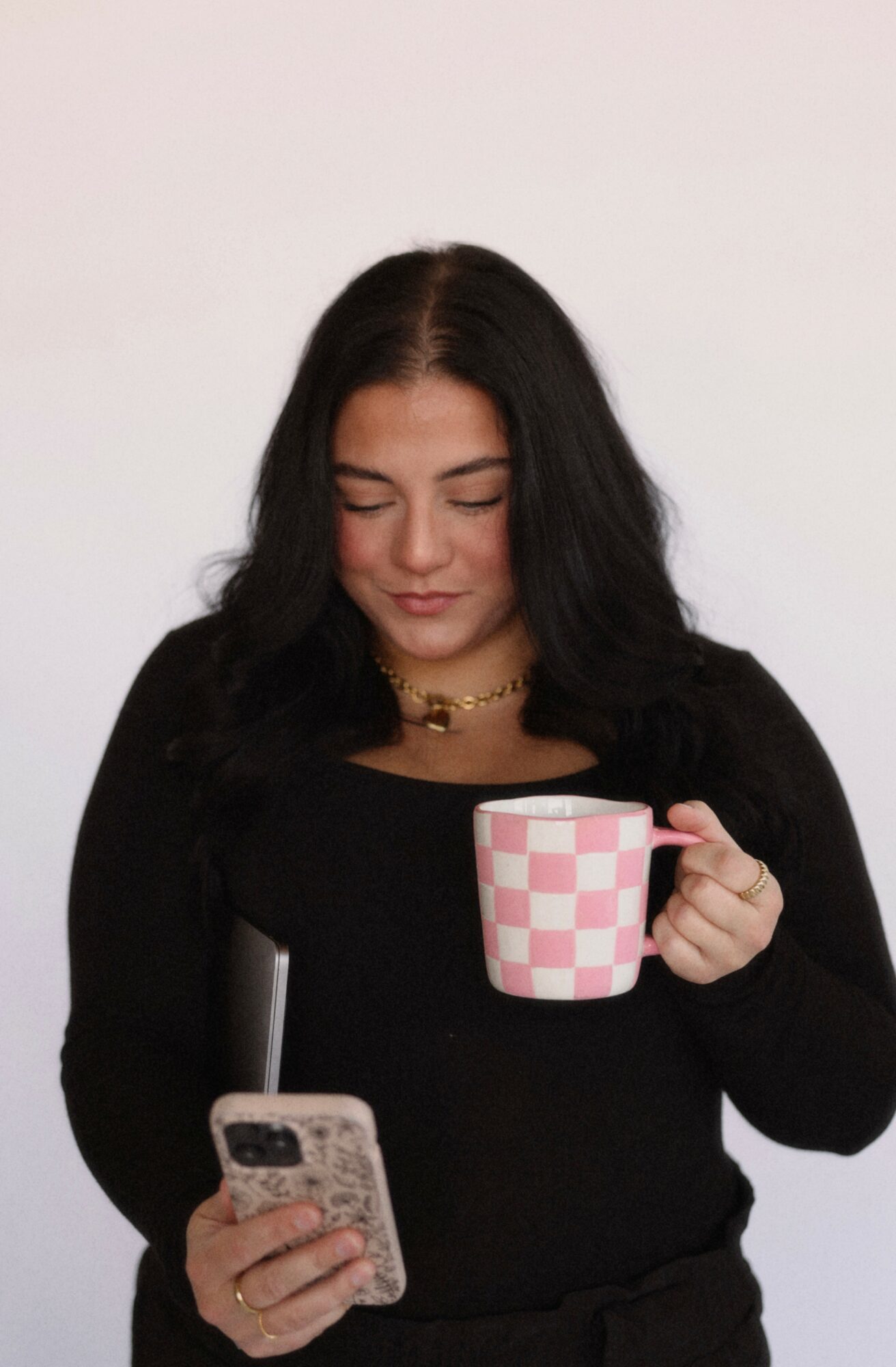 Woman with dark hair holding a checkered mug and looking at her phone, wearing a black top and necklace.