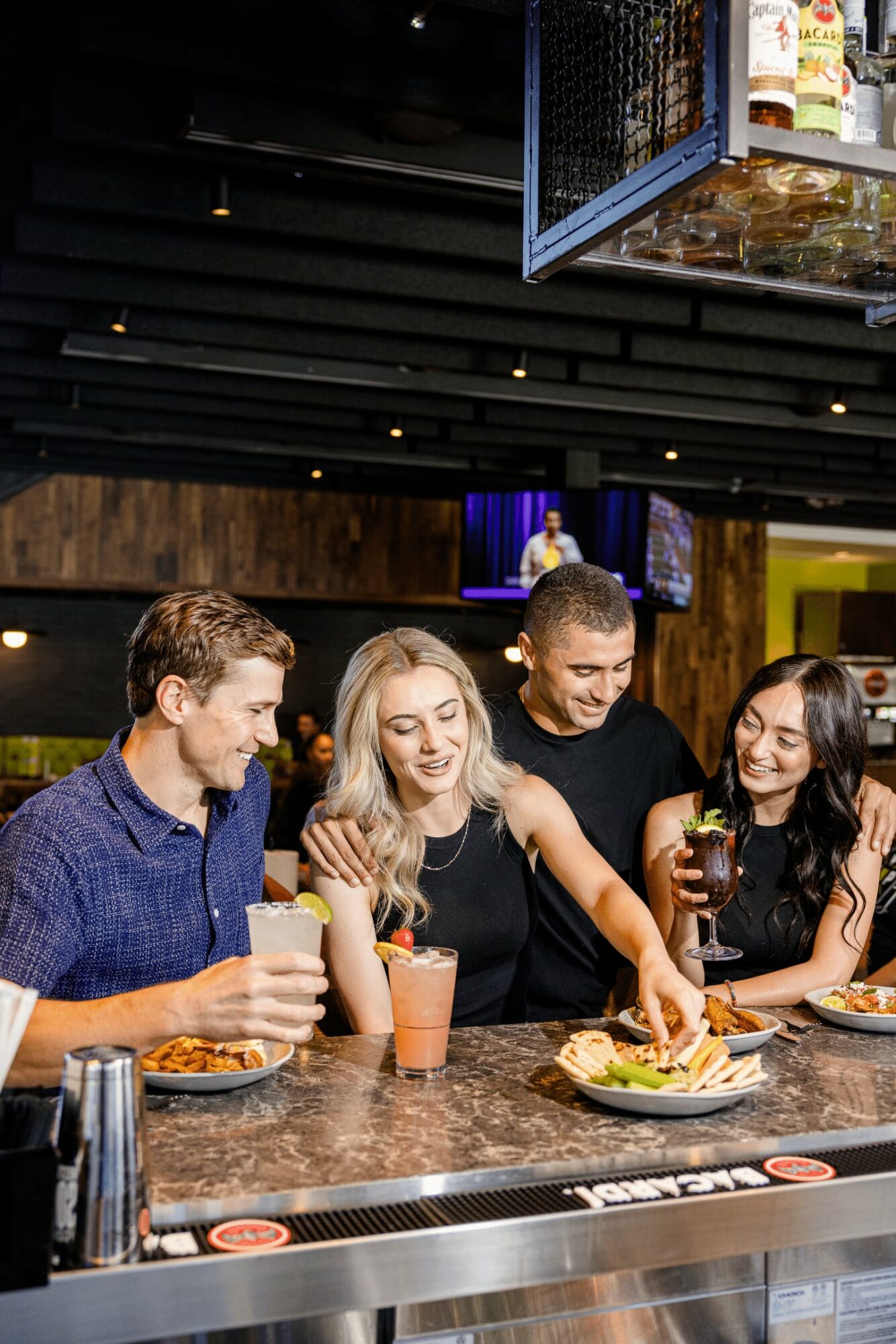 Four people sitting at a bar counter, enjoying drinks and food, with a dark ceiling and TV screens in the background.