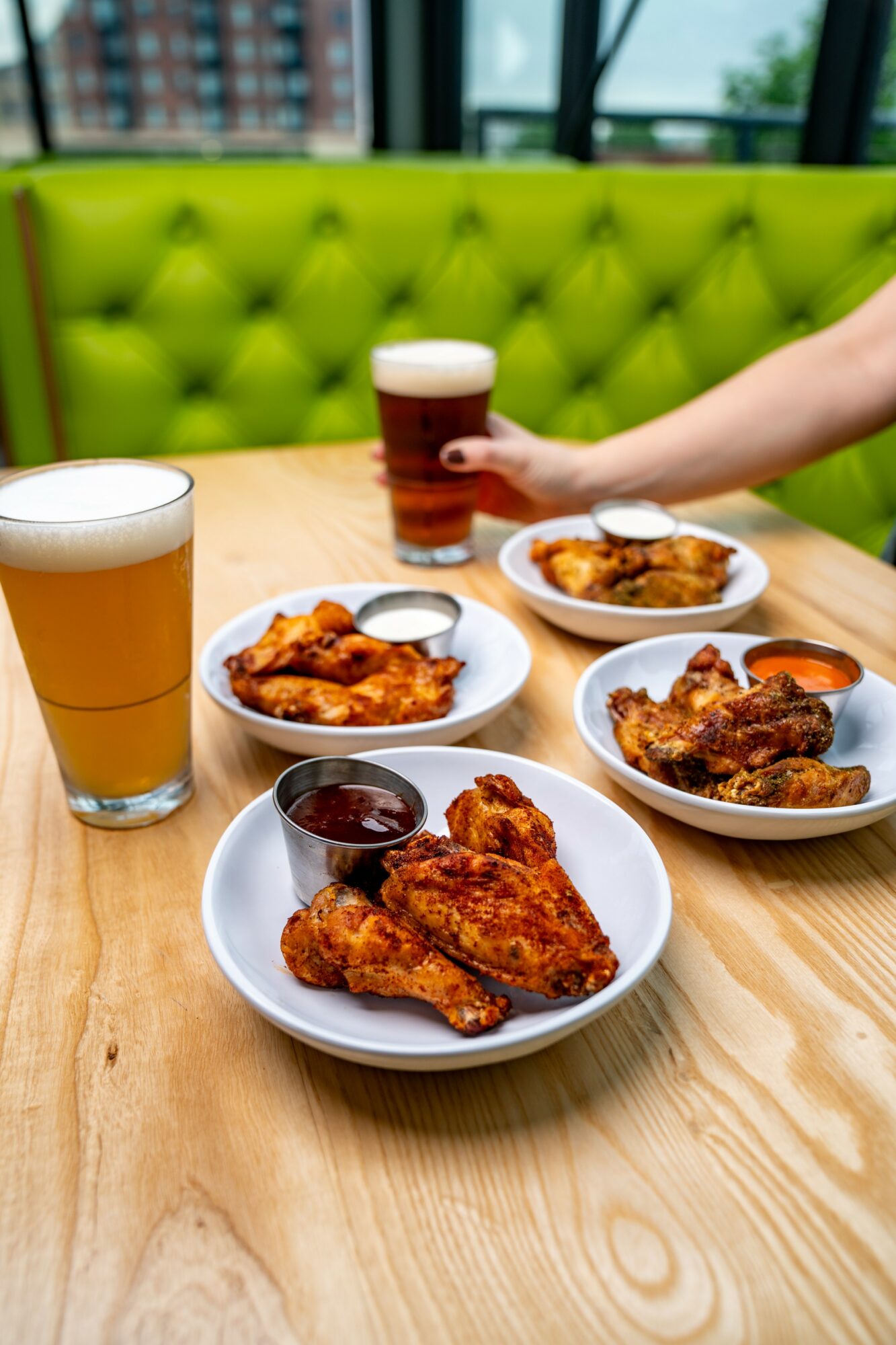 Table with three plates of fried chicken, two glasses of beer, and a hand holding a glass of beer, in a restaurant with green seating.