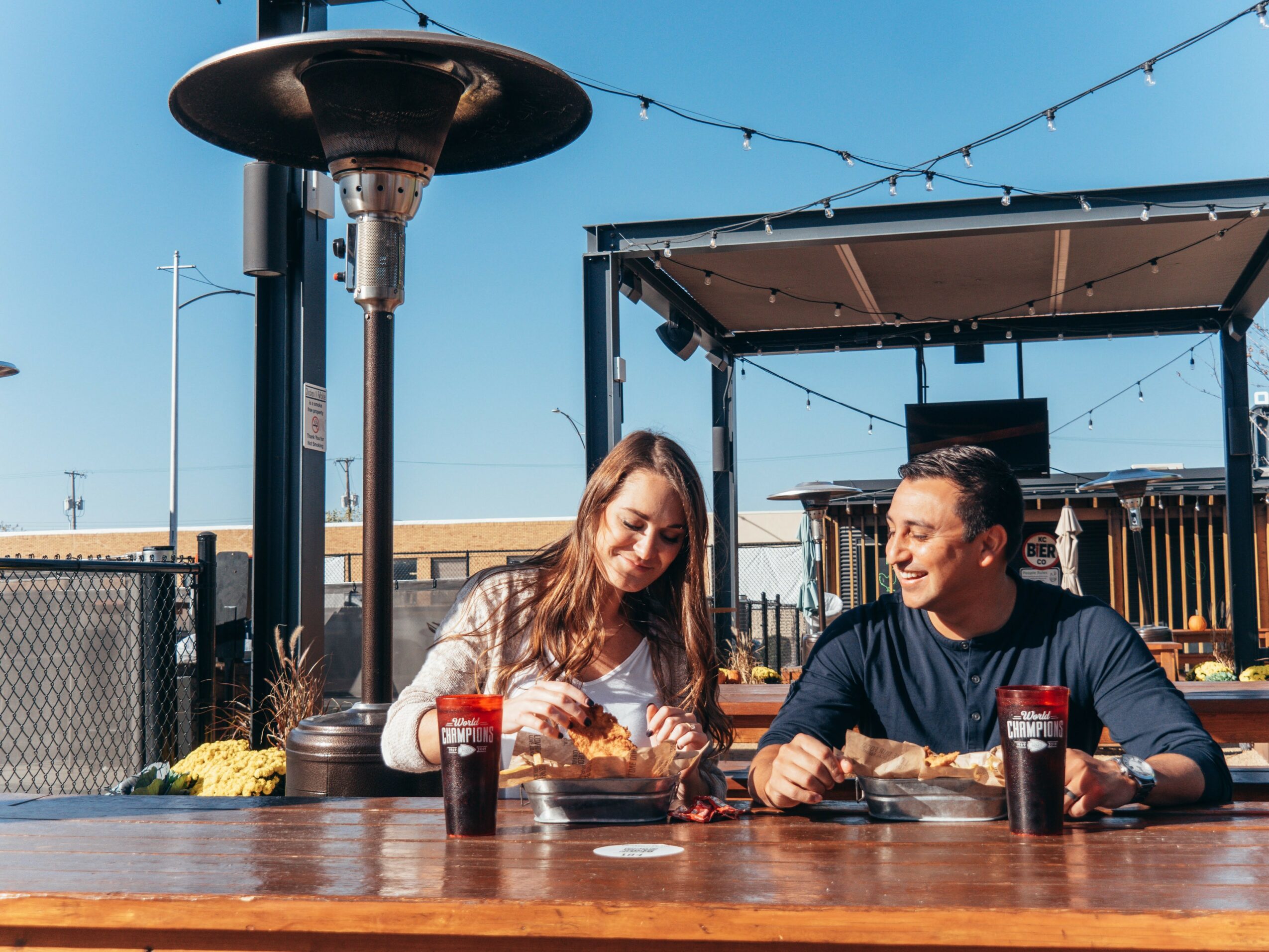 Two people sit at a wooden table outdoors, smiling and talking, with drinks in front of them, under a clear blue sky.